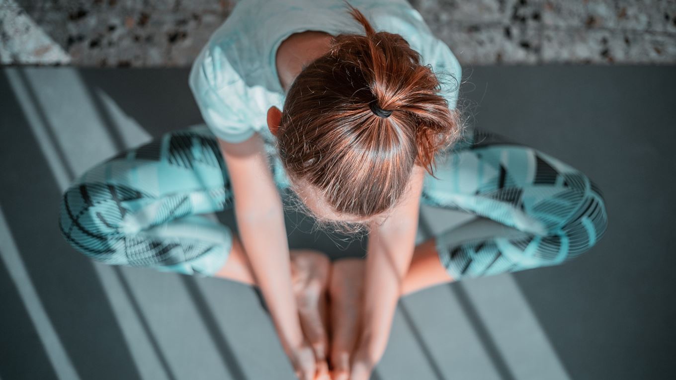 Vrouw zit in kikkerhouding op de grond. Ze zit op een grijze yogamat en kijkt naar beneden