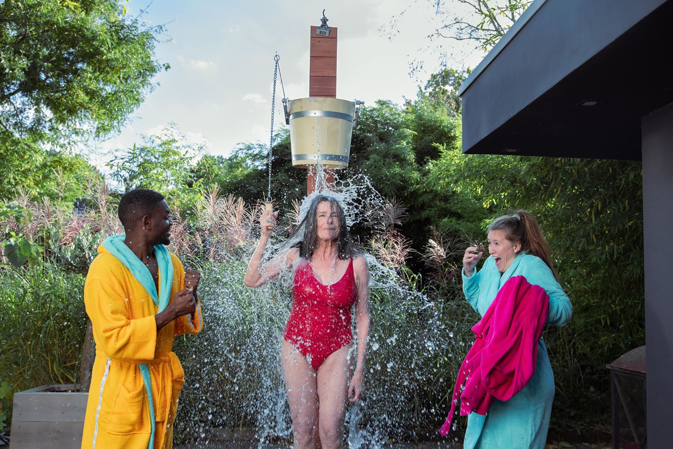 Vrouw in rood badpak staat onder een emmer met koud water. Twee mensen staan aan weerzijde van haar en kijken toe, de vrouw rechts reageert op het koude water dat zij voelt