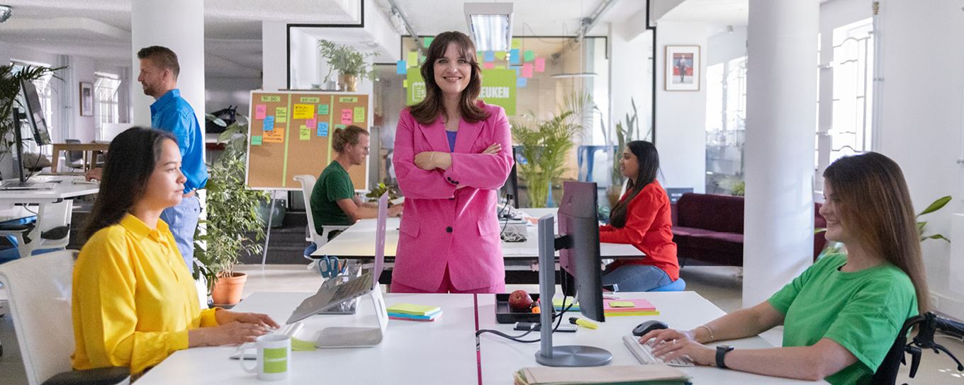 Mensen met gekleurde kleding zitten achter hun bureau. Een vrouw met roze pak staat met armen in zij en kijkt lachend in de camera