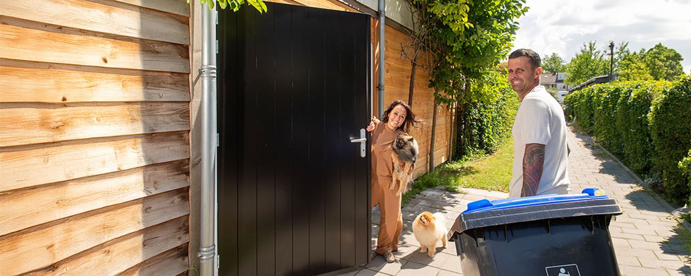 Vrouw staat bij de poort met haar hondjes en de man komt eraan met de blauwe kliko. ze lachen beiden naar de camera