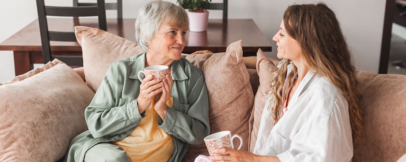 Oudere vrouw en jongere vrouw zitten samen gezellig op de bank met een kopje koffie of thee