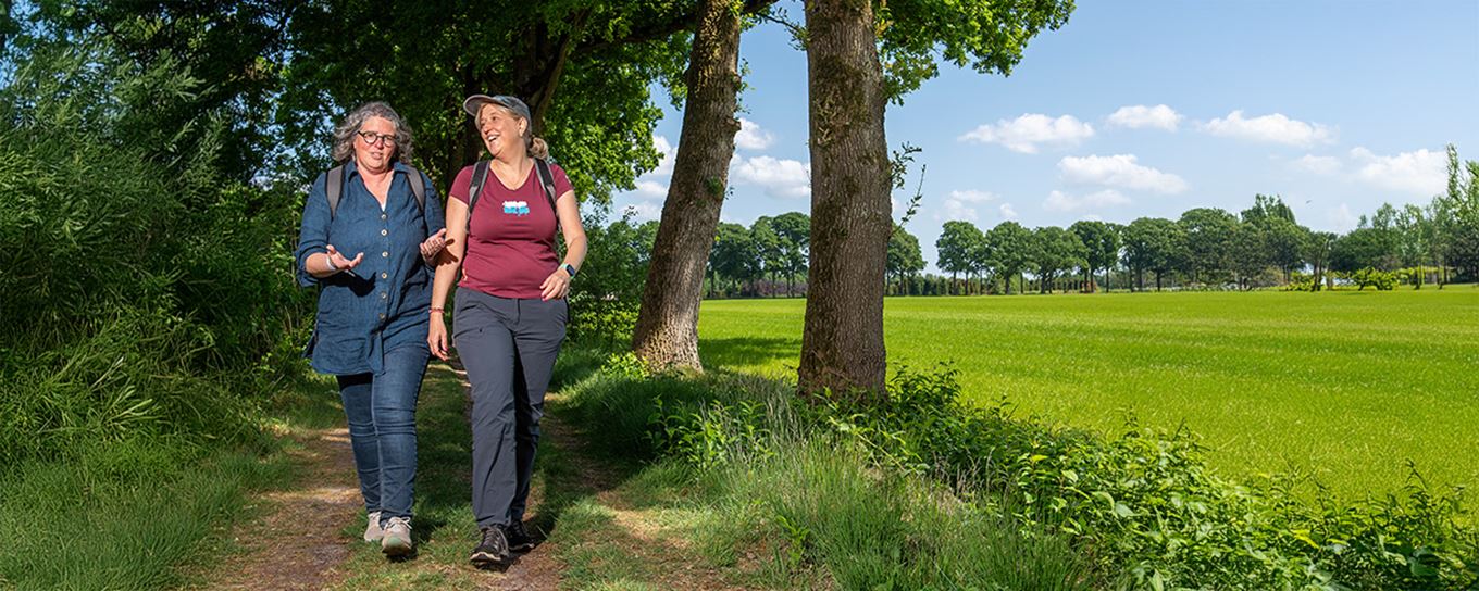 2 vrouwen van middelbare leeftijd wandelen over een wandelpad met aan de ene kant bos en aan de andere kant weiland